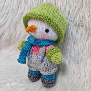 May include: A small knitted snowman toy wearing a green hat, blue scarf, and grey overalls with blue buttons. The snowman has a white body, orange nose, and black eyes.