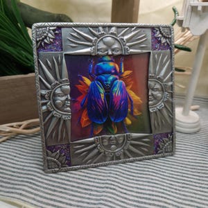 May include: A square, silver-colored metal frame with a colorful image of a beetle on a flower. The frame has sun and face designs on the corners and edges, with purple glitter accents. The beetle has iridescent blue, purple, and pink wings.