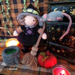 May include: A knitted witch doll with a green hat, purple dress, and broom. The doll is holding a broom and is standing next to a knitted mouse and a knitted pumpkin. The doll is on a plaid background with a candle and a cauldron.