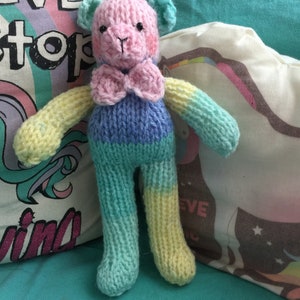 May include: A knitted teddy bear with a pink bow tie. The bear is a rainbow color with yellow, green, and blue stripes. The bear is sitting on a white pillow with a rainbow and the words "Never Stop Dreaming".