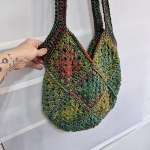 May include: A handmade crochet bag in shades of green, yellow, red, and blue. The bag features a geometric pattern and long straps. The bag is held by a hand.