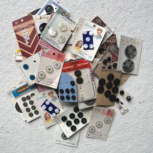 May include: A collection of vintage sewing buttons on original cardboard display cards. The assortment includes various shapes, sizes, and colors, such as white, black, and grey, with some cards featuring brand names like Penimaid, Le Chic, and Costumakers.