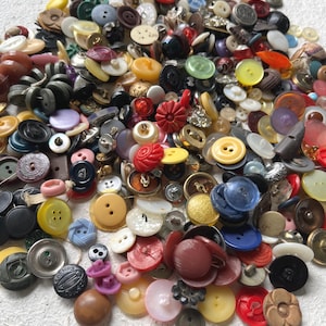 May include: A large collection of assorted sewing buttons in various shapes, sizes, and colors, including red, blue, yellow, black, white, and brown. The buttons feature different materials, textures, and hole configurations, scattered together.