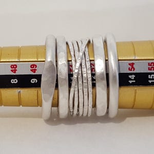 May include: A set of five silver-toned rings positioned on a yellow ring mandrel. The rings feature varied textures, including smooth, hammered, and thin crisscrossed wire designs, with the mandrel showing numerical sizing markings.