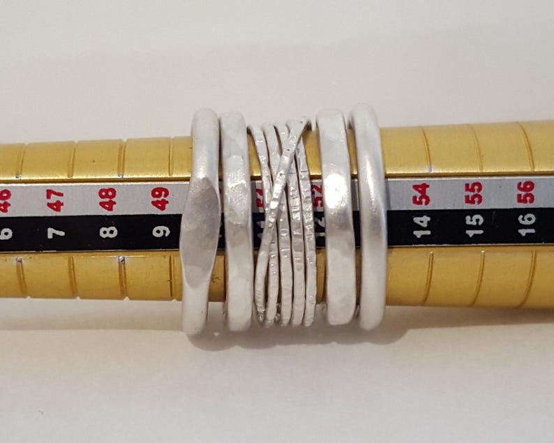 May include: A set of five silver-toned rings positioned on a yellow ring mandrel. The rings feature varied textures, including smooth, hammered, and thin crisscrossed wire designs, with the mandrel showing numerical sizing markings.
