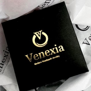May include: A square black jewelry box featuring a gold foil logo with a stylized letter V inside a circle. Below the logo, the text reads Venexia in a serif font, followed by the smaller text Modern Handmade Jewelry in a sans-serif font.