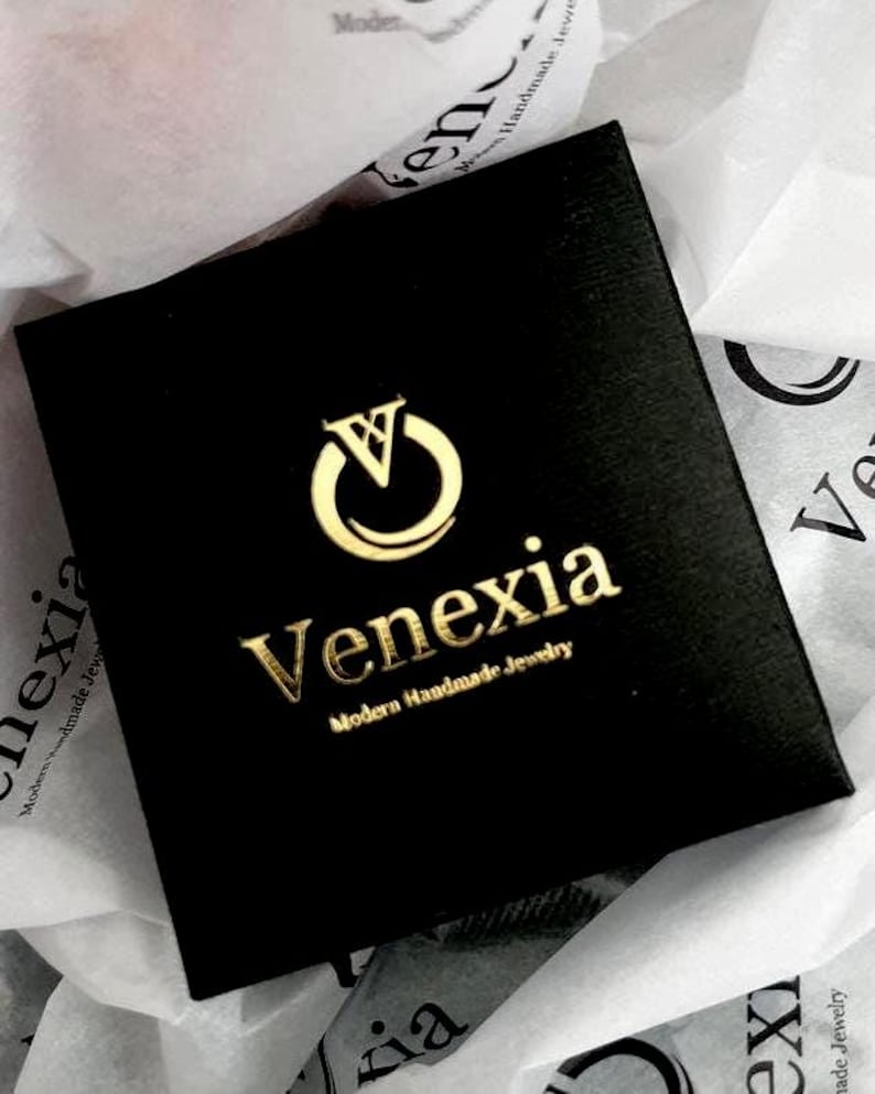 May include: A square black jewelry box featuring a gold foil logo with a stylized letter V inside a circle. Below the logo, the text reads Venexia in a serif font, followed by the smaller text Modern Handmade Jewelry in a sans-serif font.