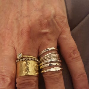 May include: A gold-toned bangle bracelet and a collection of rings. The ring finger features a wide hammered gold band, a thin band with embedded stones, and a solitaire ring. The middle finger displays a stack of five thin silver-toned bands.