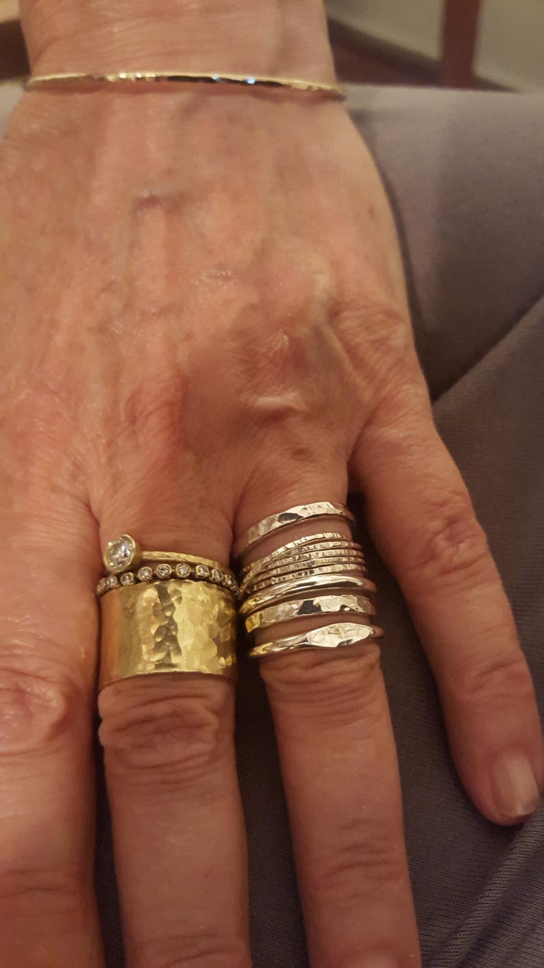 May include: A gold-toned bangle bracelet and a collection of rings. The ring finger features a wide hammered gold band, a thin band with embedded stones, and a solitaire ring. The middle finger displays a stack of five thin silver-toned bands.