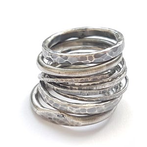 May include: A stack of seven silver-toned rings with a hammered, textured finish. Each ring features a slightly irregular, rounded band with a darkened, oxidized appearance in the indentations, creating a layered, metallic, and rustic look.