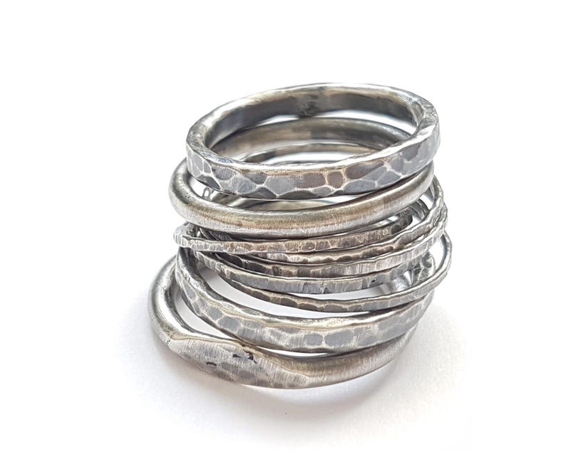 May include: A stack of seven silver-toned rings with a hammered, textured finish. Each ring features a slightly irregular, rounded band with a darkened, oxidized appearance in the indentations, creating a layered, metallic, and rustic look.