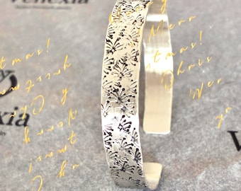 May include: A silver cuff bracelet with a floral pattern. The bracelet is open and has a textured surface. The word "Venexia" is visible in the background.