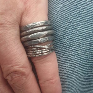 May include: A stack of five silver-toned metal rings featuring a hammered, textured finish. The rings vary in width and design, with one band displaying a smooth, elongated oval center section and another featuring a crisscross pattern.