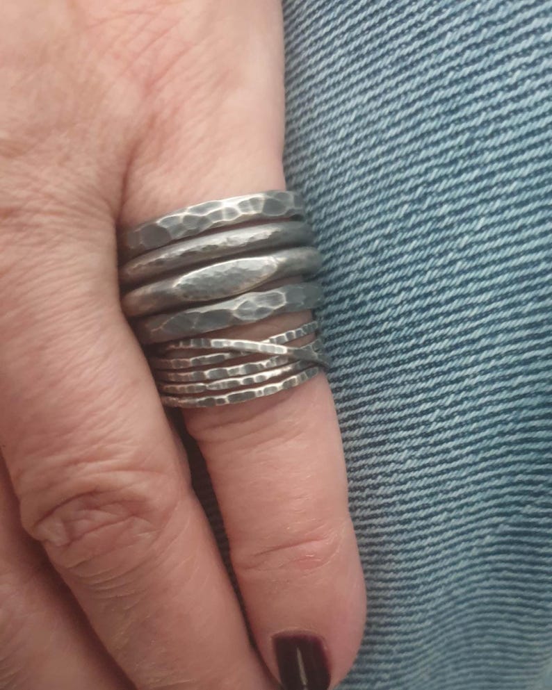 May include: A stack of five silver-toned metal rings featuring a hammered, textured finish. The rings vary in width and design, with one band displaying a smooth, elongated oval center section and another featuring a crisscross pattern.