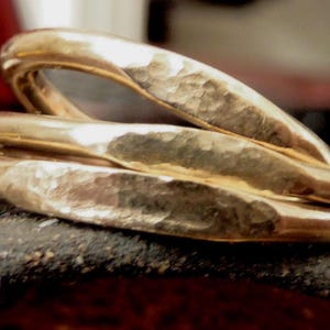 May include: Three gold-colored rings with a hammered texture are stacked together. The rings are slightly curved and have a warm, metallic sheen. The rings are resting on a dark, textured surface.