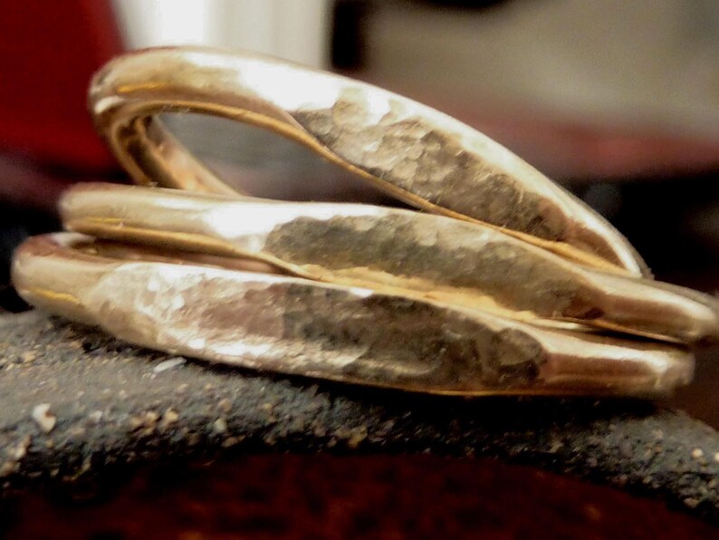 May include: Three gold-colored rings with a hammered texture are stacked together. The rings are slightly curved and have a warm, metallic sheen. The rings are resting on a dark, textured surface.