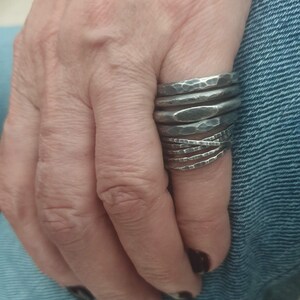 May include: A stack of multiple dark silver-toned metal rings worn on a finger. Each ring features a distinct hammered, textured surface with varying widths and shapes, creating a layered appearance across the finger from the knuckle downward.