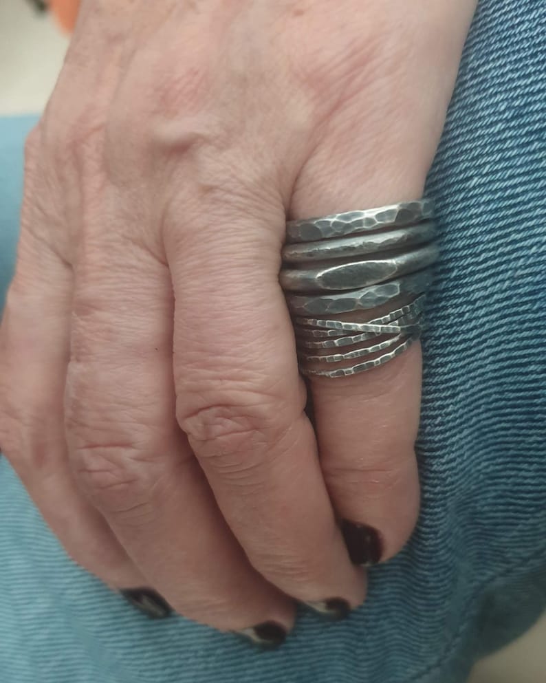 May include: A stack of multiple dark silver-toned metal rings worn on a finger. Each ring features a distinct hammered, textured surface with varying widths and shapes, creating a layered appearance across the finger from the knuckle downward.
