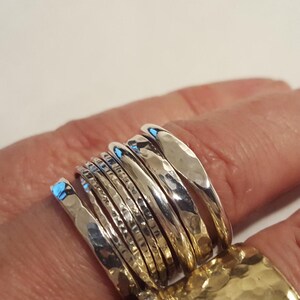 May include: A set of seven stacked silver rings featuring a variety of textures, including smooth, hammered, and faceted finishes. The rings are arranged in a row, showcasing reflective metallic surfaces and distinct patterns on each individual band.