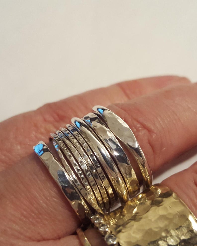 May include: A set of seven stacked silver rings featuring a variety of textures, including smooth, hammered, and faceted finishes. The rings are arranged in a row, showcasing reflective metallic surfaces and distinct patterns on each individual band.