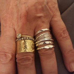 May include: A collection of rings worn on two fingers. The left finger features a wide, hammered gold band topped with a thin diamond-encrusted band and a single solitaire diamond ring. The right finger displays a stack of seven silver bands.