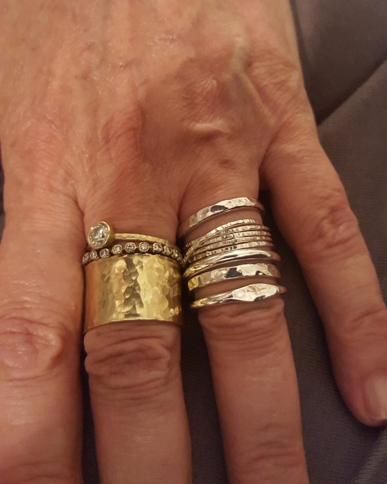 May include: A collection of rings worn on two fingers. The left finger features a wide, hammered gold band topped with a thin diamond-encrusted band and a single solitaire diamond ring. The right finger displays a stack of seven silver bands.