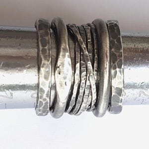 May include: A set of seven stacked silver rings featuring varied textures, including hammered surfaces, smooth rounded bands, and thin wire-like bands, all displayed on a cylindrical metal ring mandrel for sizing and display purposes.