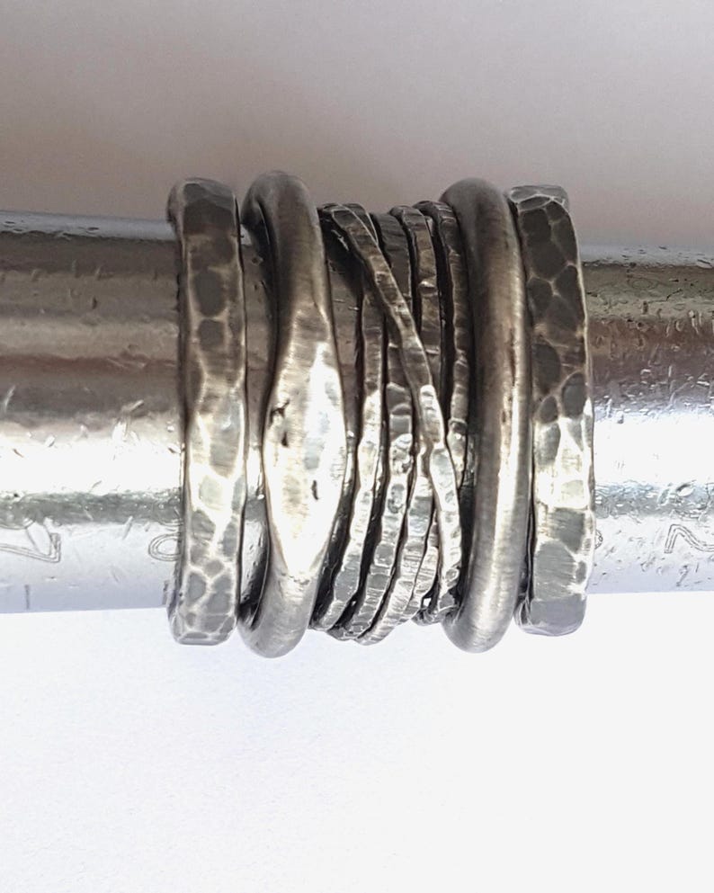May include: A set of seven stacked silver rings featuring varied textures, including hammered surfaces, smooth rounded bands, and thin wire-like bands, all displayed on a cylindrical metal ring mandrel for sizing and display purposes.