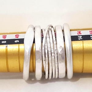 May include: A set of six silver-toned rings with varying textures and widths, positioned on a gold-colored ring mandrel. The mandrel displays numerical size markings, including 8, 9, 14, 48, 49, and 54, visible behind the stacked metal bands.