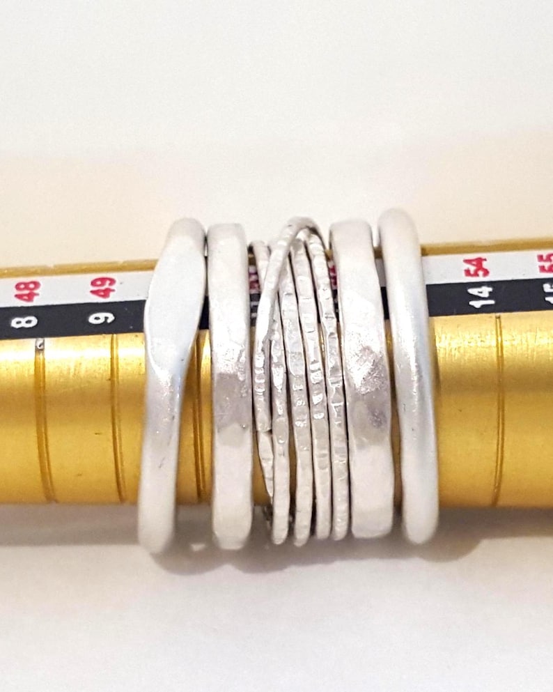 May include: A set of six silver-toned rings with varying textures and widths, positioned on a gold-colored ring mandrel. The mandrel displays numerical size markings, including 8, 9, 14, 48, 49, and 54, visible behind the stacked metal bands.