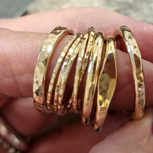 May include: A collection of gold-colored rings with hammered textures. The rings vary in width and shape, creating a layered effect. The rings are displayed on a person's finger, showcasing their reflective surfaces.