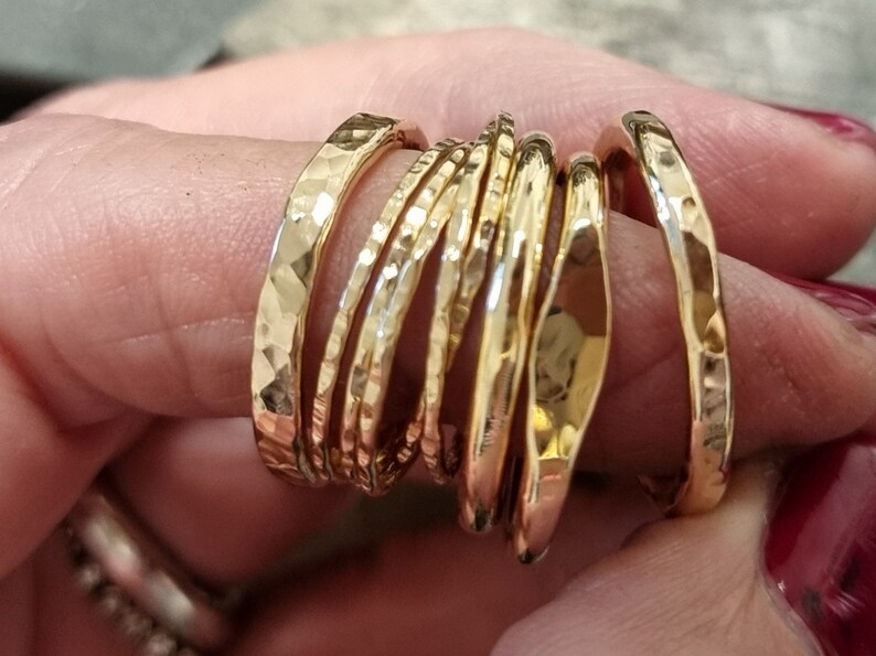 May include: A collection of gold-colored rings with hammered textures. The rings vary in width and shape, creating a layered effect. The rings are displayed on a person's finger, showcasing their reflective surfaces.