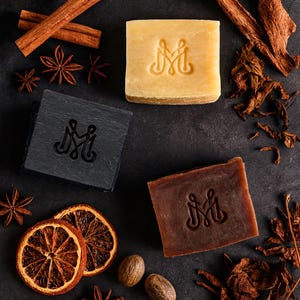 May include: Three square soaps in shades of yellow, black, and brown, each stamped with a decorative "M" design. The soaps are surrounded by cinnamon sticks, star anise, dried orange slices, and nutmeg on a dark surface.