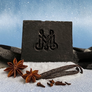 May include: A dark gray soap bar with an embossed design of two figures holding hands. The soap is surrounded by star anise, vanilla beans, and cloves on a snowy surface. A plaid scarf is in the background.