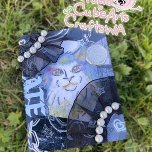 May include: A handmade journal featuring a cat illustration. The cover is decorated with black mesh, faux pearls, and a wax seal. The journal has a unique, artistic design with the text "ATE" visible.