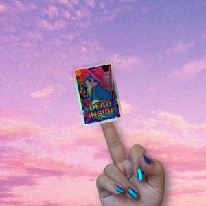 以下が含まれることがあります： ピンクの帽子をかぶった青い猫が描かれたホログラフィックステッカー。緑と青で「DEAD INSIDE」と書かれています。ステッカーはピンクと紫の空を背景に持っています。