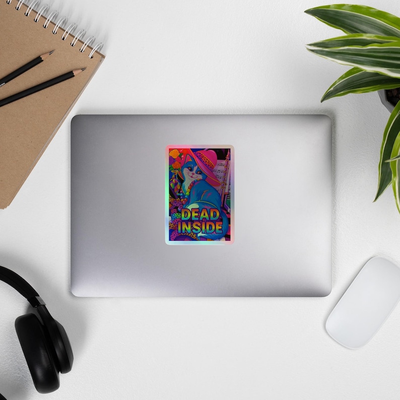 以下が含まれることがあります： ピンクの帽子をかぶった青い猫が、カラフルな花に囲まれたホログラフィックステッカー。ステッカーには「DEAD INSIDE」という文字が緑と黄色で書かれています。ステッカーはノートパソコンに貼られています。