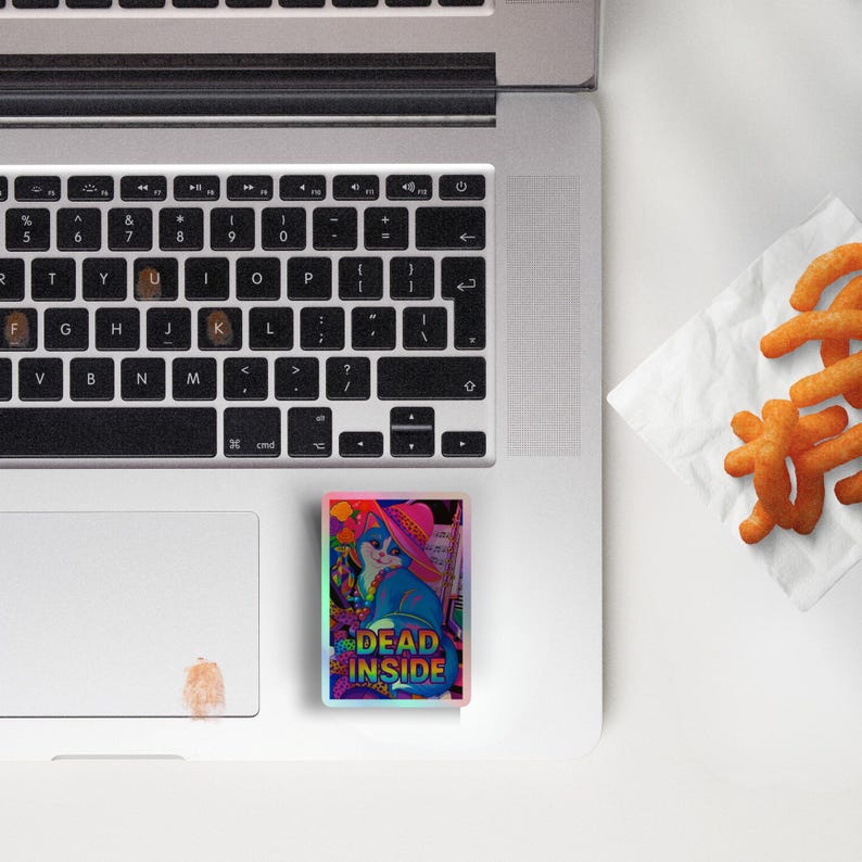 以下が含まれることがあります： ピンクの帽子をかぶった猫のカラフルなイラストと「DEAD INSIDE」の文字が入った長方形のステッカー。 ステッカーはラップトップの上にあり、オレンジ色のスナックの山に隣接しています。