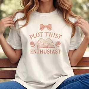 May include: Cream-colored t-shirt with the text "PLOT TWIST ENTHUSIAST" in a soft pink font. The design includes a bow, an open book, and roses. The shirt is a casual wear item.