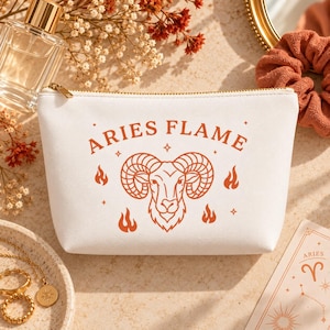 May include: A white zippered pouch featuring an orange graphic of a ram head centered between the text ARIES FLAME. The design includes small flame icons and star accents surrounding the ram. The pouch has a gold-toned metal zipper pull.
