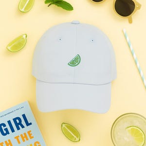 May include: A light blue baseball cap with a green lime slice embroidered on the front. The image also includes yellow sunglasses, lime slices, mint leaves, a book, a drink, and a straw.
