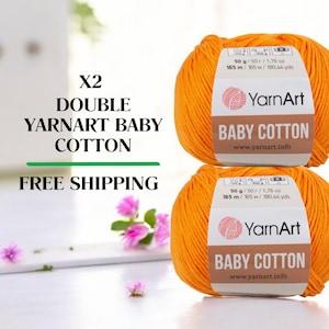 May include: Two orange YarnArt Baby Cotton yarn balls, each labeled with weight and length details. The image includes text that reads "X2 DOUBLE YARNART BABY COTTON" and "FREE SHIPPING". The yarn is displayed on a white surface with floral accents.