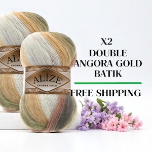 May include: Two skeins of Alize Angora Gold Batik yarn in a blend of colours including white, beige, green, and brown. The yarn is labelled with the brand name and product details. A small bouquet of purple and pink flowers is in the foreground.