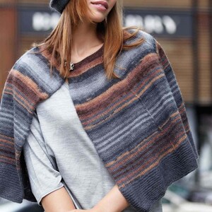 May include: A knitted cape in shades of gray, brown, and orange. The cape has horizontal stripes and is worn over a gray top. A matching gray hat is also visible. The cape is a stylish accessory for cooler weather.