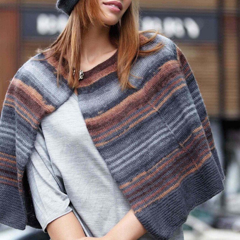 May include: A knitted cape in shades of gray, brown, and orange. The cape has horizontal stripes and is worn over a gray top. A matching gray hat is also visible. The cape is a stylish accessory for cooler weather.