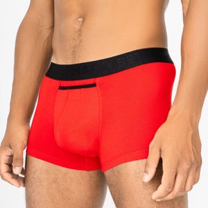 Puede incluir: Bóxer rojo brillante con cintura negra. La cintura muestra el texto "MAN'S SET". El bóxer tiene un diseño de bolsillo frontal y un detalle de ribete negro.