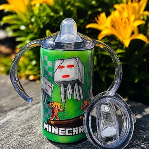 K&ouml;nnte beinhalten: Ein gr&uuml;ner, glitzernder Schnabelbecher mit zwei transparenten Griffen und einem durchsichtigen Deckel. Der Becher zeigt ein Minecraft-Motiv mit einem wei&szlig;en Ghast, einer Figur und dem Schriftzug MINECRAFT. Ein separater Deckel liegt daneben.