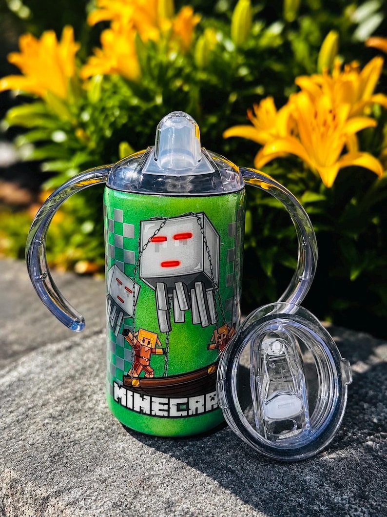 K&ouml;nnte beinhalten: Ein gr&uuml;ner, glitzernder Schnabelbecher mit zwei transparenten Griffen und einem durchsichtigen Deckel. Der Becher zeigt ein Minecraft-Motiv mit einem wei&szlig;en Ghast, einer Figur und dem Schriftzug MINECRAFT. Ein separater Deckel liegt daneben.