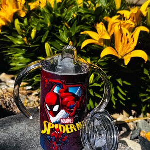 K&ouml;nnte beinhalten: Ein zylindrischer Schnabelbecher mit zwei transparenten Kunststoffgriffen und einem durchsichtigen Deckel. Der Becher zeigt eine rote und blaue Marvel Spider-Man-Grafik mit dem gelb gedruckten Text Spider-Man. Das Design enth&auml;lt eine gro&szlig;e Figur.