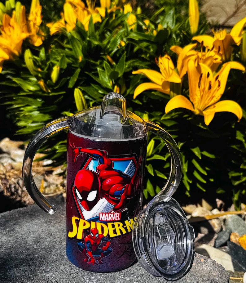 K&ouml;nnte beinhalten: Ein zylindrischer Schnabelbecher mit zwei transparenten Kunststoffgriffen und einem durchsichtigen Deckel. Der Becher zeigt eine rote und blaue Marvel Spider-Man-Grafik mit dem gelb gedruckten Text Spider-Man. Das Design enth&auml;lt eine gro&szlig;e Figur.